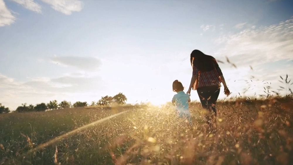 Mom holding hands with her daughter and walking in a field towards the sunset.